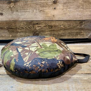 May include: A round, camouflage-patterned cushion with a black border and a black handle. The cushion features a mix of brown, green, and tan colors, creating a natural, outdoor aesthetic. The cushion is designed for comfort and portability.