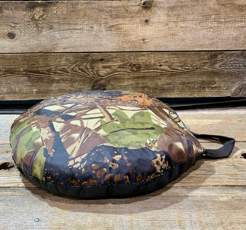 May include: A round, camouflage-patterned cushion with a black border and a black handle. The cushion features a mix of brown, green, and tan colors, creating a natural, outdoor aesthetic. The cushion is designed for comfort and portability.