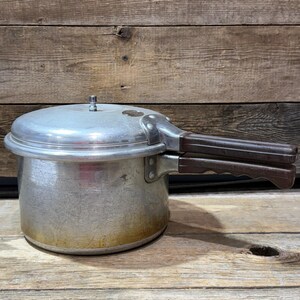 Puede incluir: Una olla a presión plateada vintage con asas marrones. La olla tiene un cuerpo redondo y una tapa con una válvula de alivio de presión. Las asas son de un material marrón oscuro. La olla está sobre una superficie de madera.