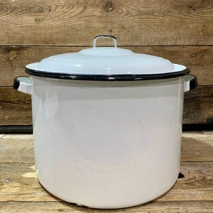 May include: A large, white enamel pot with a lid and two side handles. The pot has a black rim and handle accents. The lid has a small handle. The pot is speckled and sits on a wooden surface.