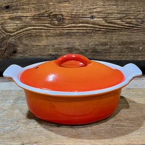 Pode incluir: Uma panela de ferro fundido esmaltado laranja com tampa. A tampa tem uma alça vermelha e a borda é branca. A panela tem alças brancas em ambos os lados. O fundo é uma superfície de madeira rústica.