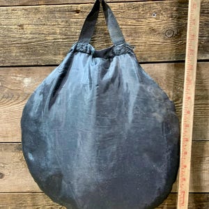 May include: A dark gray, round, drawstring bag with a black handle. The bag is made of a shiny, possibly nylon, material. A wooden ruler is placed next to the bag.
