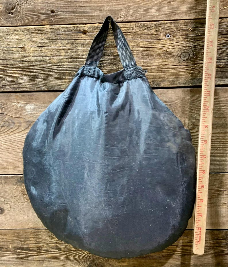 May include: A dark gray, round, drawstring bag with a black handle. The bag is made of a shiny, possibly nylon, material. A wooden ruler is placed next to the bag.
