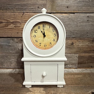 May include: A white wooden tabletop clock featuring a circular face with black Arabic numerals and a distressed tan background. The clock is topped with a small finial and includes a single rectangular storage drawer with a round knob at the base.