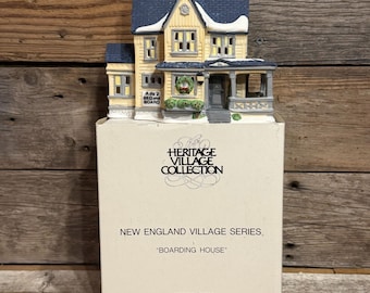 May include: A porcelain miniature house from The Heritage Village Collection New England Village Series titled Boarding House. The yellow building features a blue roof with snow accents, a front porch, a wreath, and a sign reading Ado's Bed and Board.