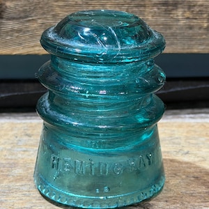 Könnte beinhalten: Ein vintage elektrischer Isolator aus türkisfarbenem Glas mit abgerundeter Oberseite und gestuften Rillen. Die Basis weist eine geprägte Beschriftung mit der Aufschrift HEMINGRAY auf. Das Objekt ist durchscheinend mit sichtbaren Luftblasen.