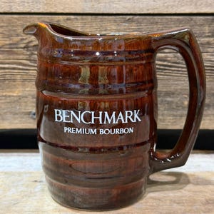 Puede incluir: Una jarra de cerámica marrón con asa y boquilla. La jarra tiene un diseño en forma de barril y las palabras "BENCHMARK PREMIUM BOURBON" en letras blancas. La jarra está esmaltada y tiene un acabado brillante.