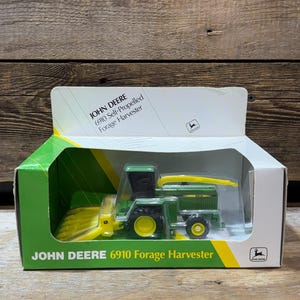Puede incluir: Modelo de juguete de la cosechadora de forraje autopropulsada John Deere 6910 en su caja original. El juguete es verde y amarillo, con detalles en negro. La caja es verde y blanca, con el logotipo de John Deere e información del producto.