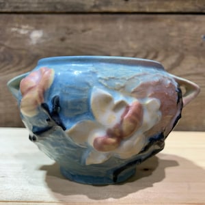 Puede incluir: Un cuenco de cerámica azul con dos asas y detalles florales en relieve. El cuenco presenta un color base azul claro con acentos rosas, crema y negros. El diseño floral es una característica destacada del cuenco.