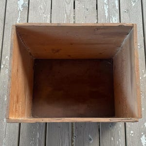 May include: A rectangular wooden box with a tapered interior. The box is constructed from light brown wood, with darker brown tones inside. The box has a rustic, aged appearance, suggesting it may be vintage or antique. The box is empty.
