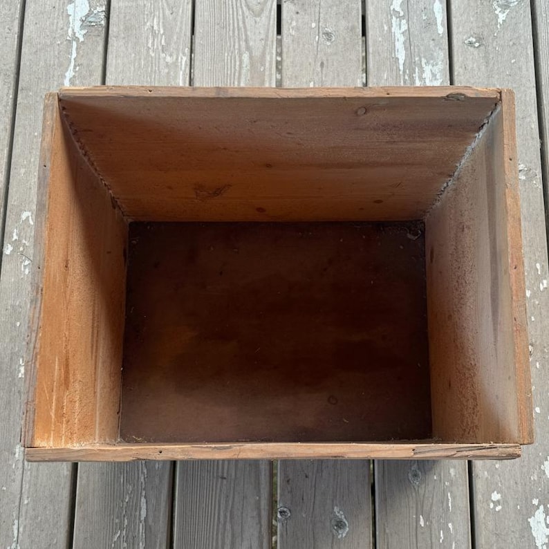 May include: A rectangular wooden box with a tapered interior. The box is constructed from light brown wood, with darker brown tones inside. The box has a rustic, aged appearance, suggesting it may be vintage or antique. The box is empty.