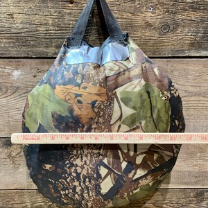 May include: A camouflage-patterned tote bag with a black handle. The bag is approximately 12 inches wide. The bag's design features shades of green, brown, and tan, creating a woodland camouflage effect. The bag is made of a lightweight material.