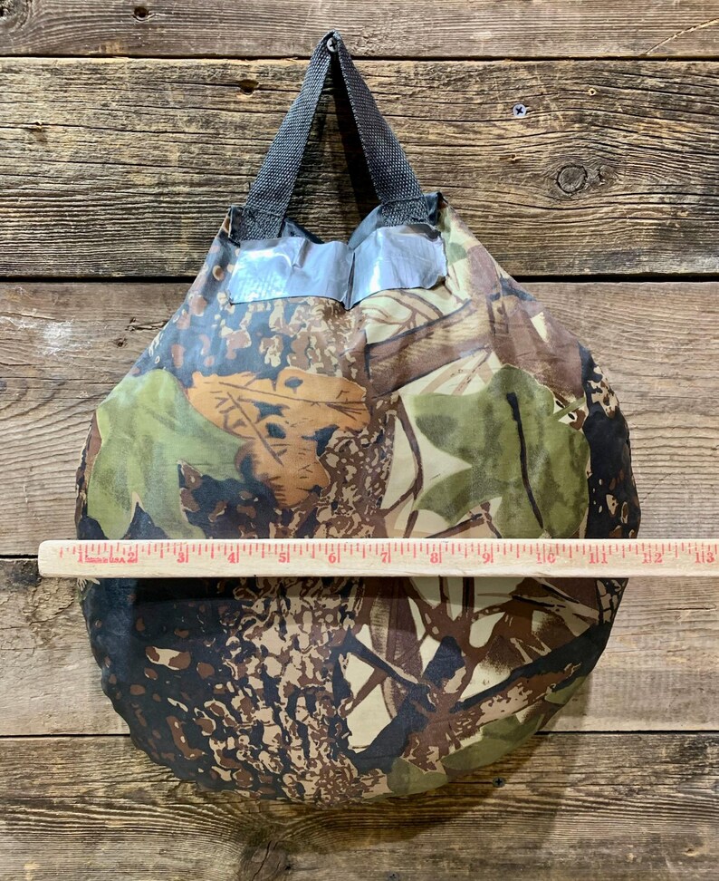 May include: A camouflage-patterned tote bag with a black handle. The bag is approximately 12 inches wide. The bag's design features shades of green, brown, and tan, creating a woodland camouflage effect. The bag is made of a lightweight material.