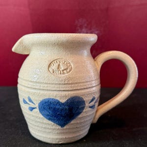 Puede incluir: Una jarrita de gres color crema con superficie texturizada, que presenta un corazón azul pintado en la parte frontal y un pequeño logotipo circular estampado cerca del borde. La jarra tiene un asa curva, un pico vertedor y estrías.