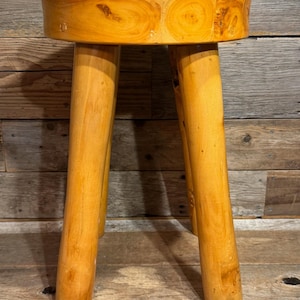 May include: A round wooden stool with a smooth, light brown finish featuring visible natural wood grain patterns. The stool is supported by three thick, cylindrical legs that extend downward from the circular seat, showcasing a polished texture.
