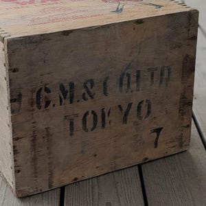 May include: A weathered wooden crate with stenciled black text reading "C.M. & CO. LTD TOKYO 7". The crate has visible wood grain and metal fasteners. The top of the crate has faded red text and a small illustration.