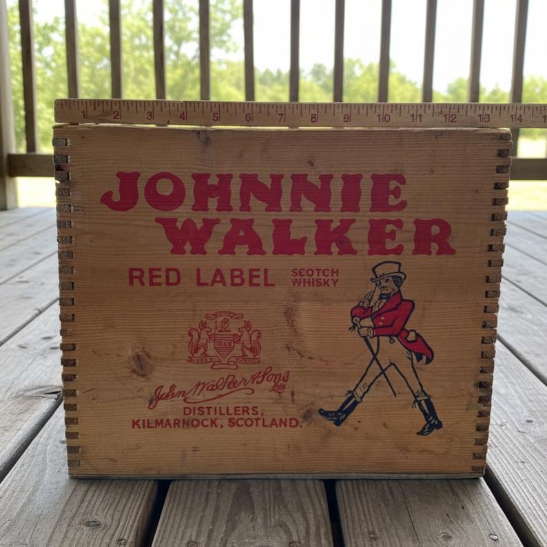 May include: Wooden crate with the words "JOHNNIE WALKER RED LABEL SCOTCH WHISKY" in red lettering. The crate features an illustration of a man in a red coat and top hat. The text "John Walker & Sons, Distillers, Kilmarnock, Scotland" is also visible.