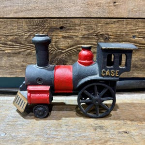 May include: A cast iron toy train painted black and red. The train features a smokestack, a cab with the word "CASE", and detailed wheels. The train is a decorative item.