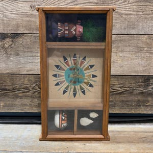 Puede incluir: Una vitrina de madera con frente de vidrio, que muestra un reloj y figuras de temática nativa americana. El reloj presenta un diseño de sol con un centro turquesa. La vitrina también contiene una pequeña muñeca y elementos decorativos.