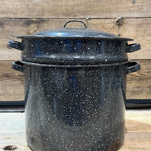 Puede incluir: Una olla de esmalte negro con tapa y asas laterales. La olla tiene un patrón blanco moteado y está sobre una superficie de madera. La olla está diseñada para conservar alimentos.