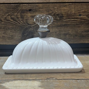 Peut inclure: Beurrier en céramique blanche avec un couvercle en forme de dôme et un bouton en verre transparent. La base rectangulaire a un bord perlé. Le couvercle a des rainures verticales. Le plat est sur une surface en bois.
