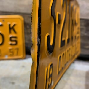 May include: Close-up of a vintage yellow license plate with black embossed numbers and letters. The plate has a distressed look with visible wear and tear. The text on the plate reads "3216 COLORADO" and "19". Another yellow plate is visible in the background.