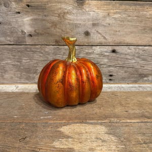 May include: A decorative pumpkin figurine featuring a vibrant orange and red gradient finish with a distressed, textured surface. The pumpkin has a prominent, upright stem finished in a bright, reflective metallic gold colour.