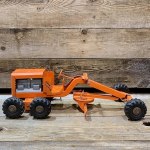 May include: An orange toy grader with black tires. The grader has a detailed design, including a steering wheel and a blade. The toy is made of metal and sits on a wooden surface.