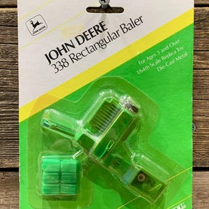Peut inclure: Un jouet John Deere 338 Rectangular Baler, en métal moulé sous pression, dans un emballage vert et jaune. L'emballage comprend le logo John Deere et le texte "For Ages 3 and Over". Le jouet est une réplique à l'échelle 1/64.