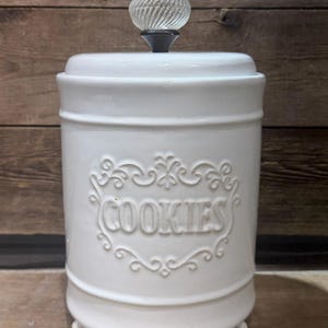 Może przedstawiać: Biały ceramiczny pojemnik na ciasteczka z ozdobną pokrywką i przezroczystym, teksturowanym pokrętłem. Słowo „COOKIES” jest wytłoczone z przodu w ozdobnym wzorze. Słoik ma cylindryczny kształt i stoi na małych nóżkach.