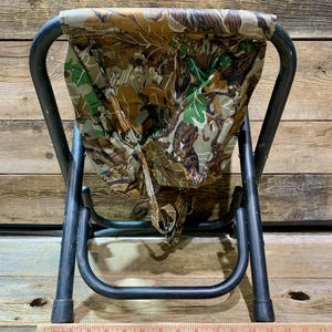 May include: A folding stool with a camouflage seat and black metal frame. The seat features a pattern of brown, green, and tan leaves. The stool is designed for outdoor use and is compact when folded.
