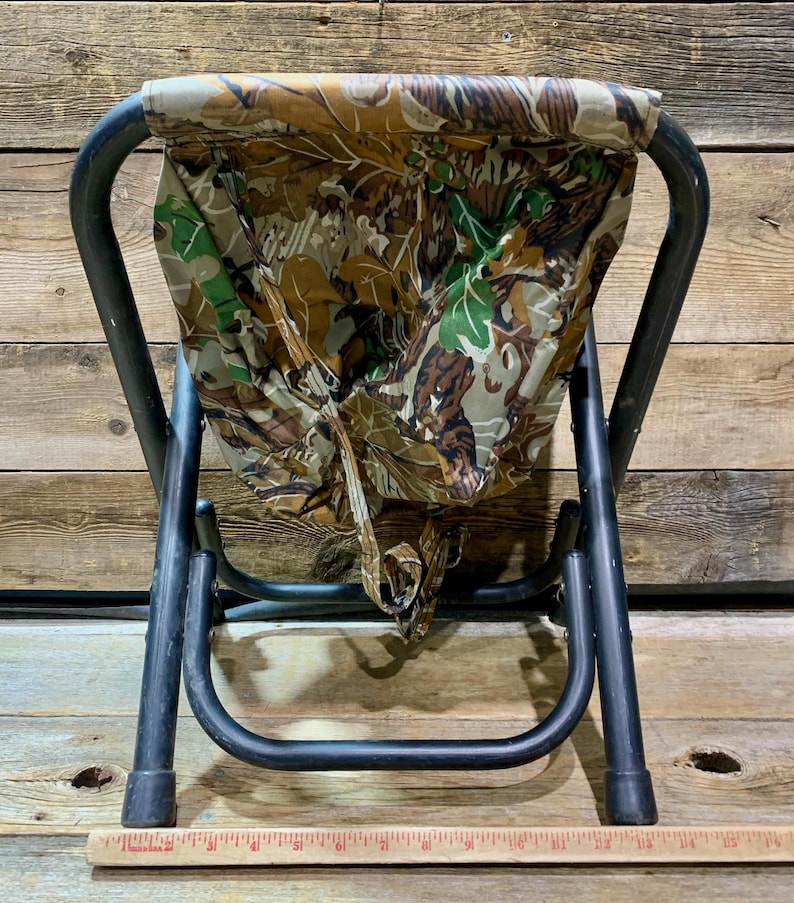 May include: A folding stool with a camouflage seat and black metal frame. The seat features a pattern of brown, green, and tan leaves. The stool is designed for outdoor use and is compact when folded.