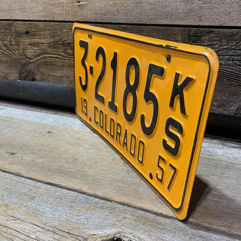 May include: A vintage Colorado license plate with black lettering and numbers on a yellow background. The plate reads "3-2185 KS 19 COLORADO 57". The plate is slightly angled, showing its aged condition and the wooden surface it rests on.