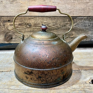 May include: Vintage copper kettle with a dark red wooden handle and lid knob. The kettle has a spout and a curved metal handle. The copper has a weathered, aged appearance, with areas of discoloration and patina, suggesting its age and use.