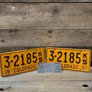 May include: Two vintage Colorado license plates with the number 3-2185. The plates are yellow with black lettering and numbers. One plate reads "19. COLORADO" and the other reads "LORADO 57". A small sign is between the plates.