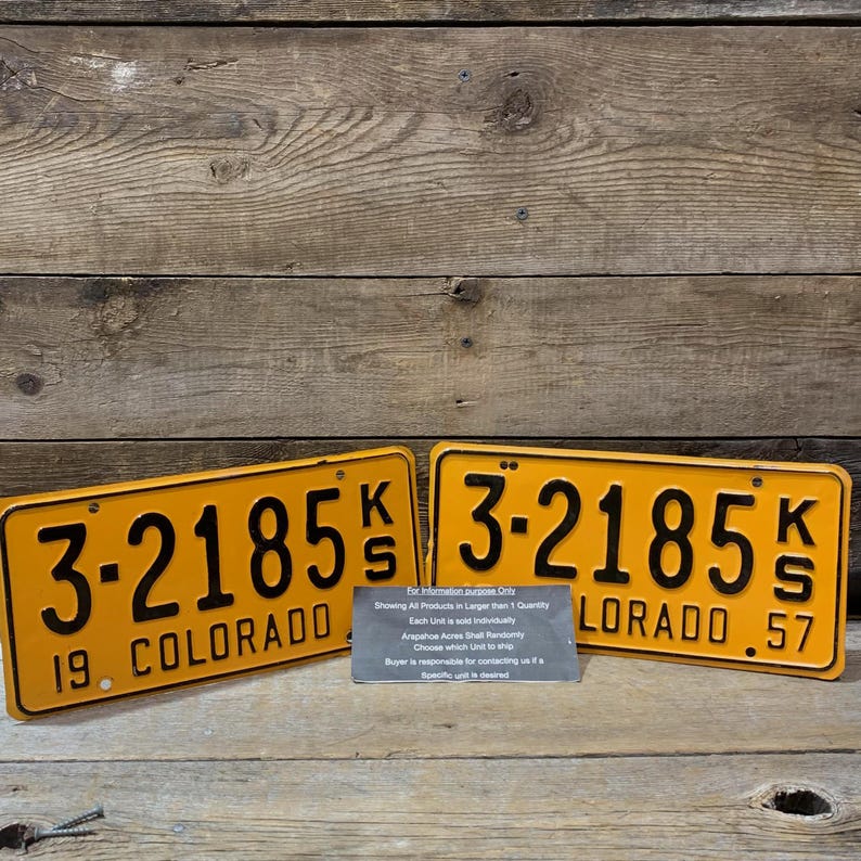 May include: Two vintage Colorado license plates with the number 3-2185. The plates are yellow with black lettering and numbers. One plate reads "19. COLORADO" and the other reads "LORADO 57". A small sign is between the plates.