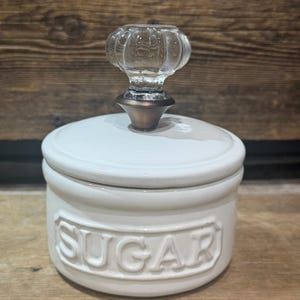 May include: A white ceramic sugar jar with a lid and a clear glass knob. The word "SUGAR" is embossed on the front of the jar. The jar sits on a wooden surface.