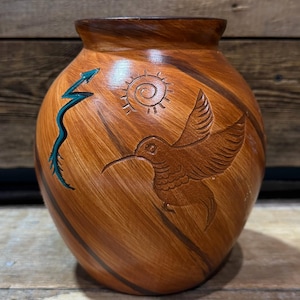 May include: A handcrafted, brown ceramic vase with a textured finish. The vase features carved designs of a hummingbird, sun, and lightning bolt. The rim and base are a darker shade of brown.