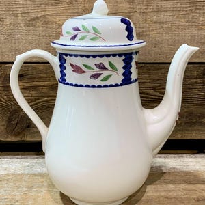 May include: White ceramic coffee pot with a lid. The pot features a curved handle and spout. The lid and upper portion of the pot are decorated with a blue border and floral designs in red, green, and purple. The pot sits on a wooden surface.