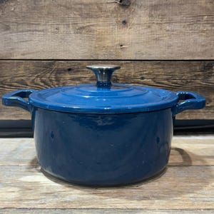 Puede incluir: Una olla holandesa de hierro fundido esmaltado azul con tapa y dos asas laterales. La tapa tiene un pomo plateado. La olla está sobre una superficie de madera.