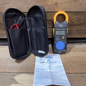 May include: A black zippered case with a clamp meter and instruction manual. The clamp meter is black and yellow, displaying a reading of 0.004. The instruction manual is white with blue text. The case is open, revealing a compartment with wires.