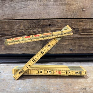 May include: A vintage wooden folding ruler, featuring brass joints and markings in black and red. The ruler is partially extended, displaying inch measurements from 1 to 19. The brand name "LUFKIN" is visible.