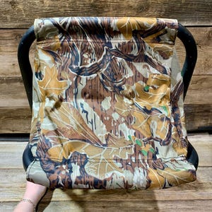 May include: A camouflage-patterned folding stool with a black frame. The fabric features a mix of brown, tan, and green leaf and branch designs. The stool has a curved handle on each side for easy carrying.