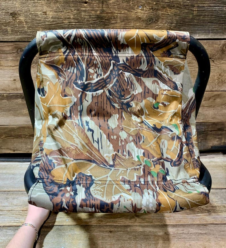 May include: A camouflage-patterned folding stool with a black frame. The fabric features a mix of brown, tan, and green leaf and branch designs. The stool has a curved handle on each side for easy carrying.