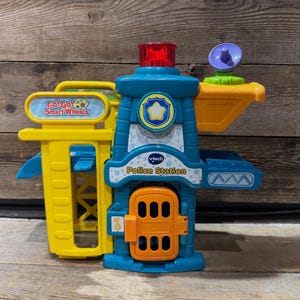 May include: A colorful VTech Go! Go! Smart Wheels police station playset. The toy features a blue tower with a red light, a yellow parking structure, and an orange door. The words "Police Station" and "Go! Go! Smart Wheels" are visible.