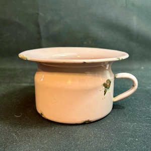 May include: A vintage, pink enamel chamber pot with a handle. The pot has a wide rim and a cylindrical body. The paint is chipped in several places, giving it a rustic appearance. The handle is small and curved.