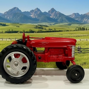 Może przedstawiać: Czerwony traktor zabawka z czarnymi oponami i srebrnymi felgami. Traktor ma klasyczny design i jest ustawiony na tle zielonego pola i gór. Koła traktora są duże, a karoseria ma jasnoczerwony kolor.