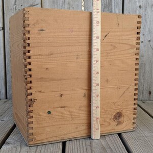 May include: A square, unfinished wooden box with dovetail joints. A wooden ruler is placed next to the box, showing a height of approximately 10 inches. The box is made of light-colored wood.