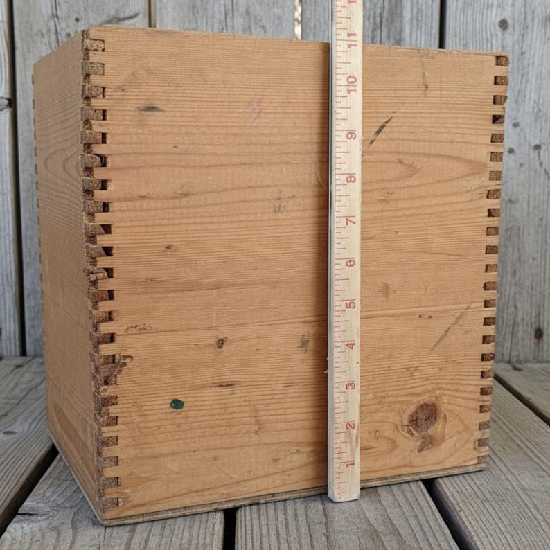 May include: A square, unfinished wooden box with dovetail joints. A wooden ruler is placed next to the box, showing a height of approximately 10 inches. The box is made of light-colored wood.