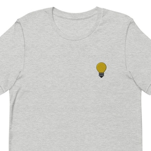 May include: A pale grey t-shirt with a small embroidered yellow lightbulb design on the left chest. The lightbulb has a black base and a yellow bulb. The shirt has short sleeves and a crew neck.